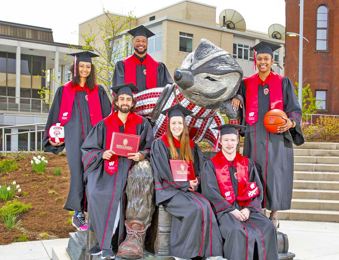Various student athletes who graduated in May of 2019 sitting in their graduation outfits around a Bucky statue.